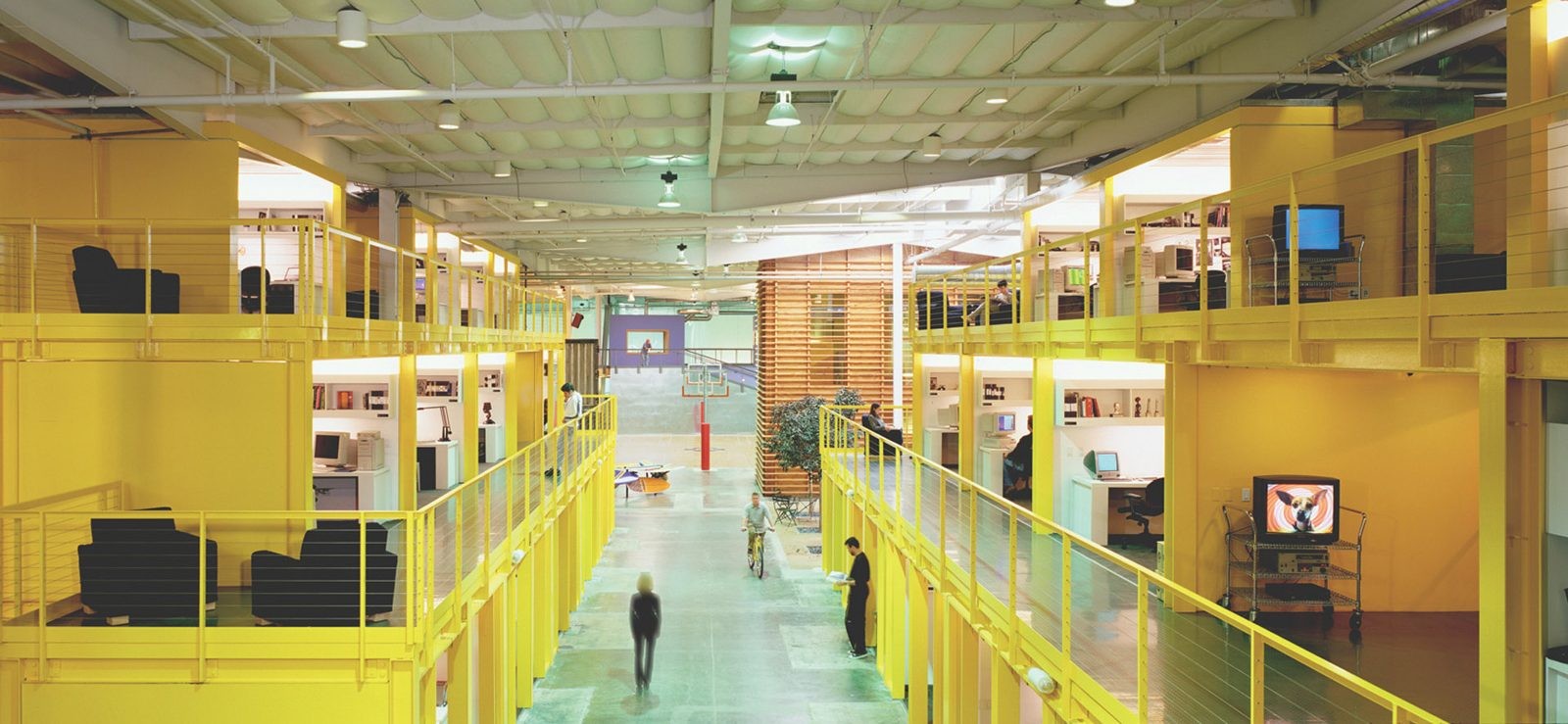 The "Main Street" at TBWA\Chiat\Day's Los Angeles office, 1998. After a failed attempt to abolish desks entirely four years prior, Clive Wilkinson redesigned the converted warehouse using urban planning principles. He created an indoor "town" featuring two-story structures lining a green floor, connected by bridges to encourage casual friction.